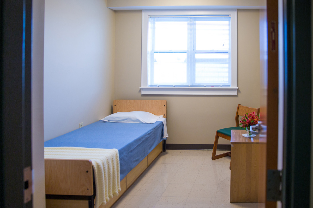 a small, unadorned room with a bed, window, dresser and chair.