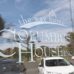 Close up of lettering on a window: "The way home, Columbus House Inc." Visible through the window are two cars and some trees.